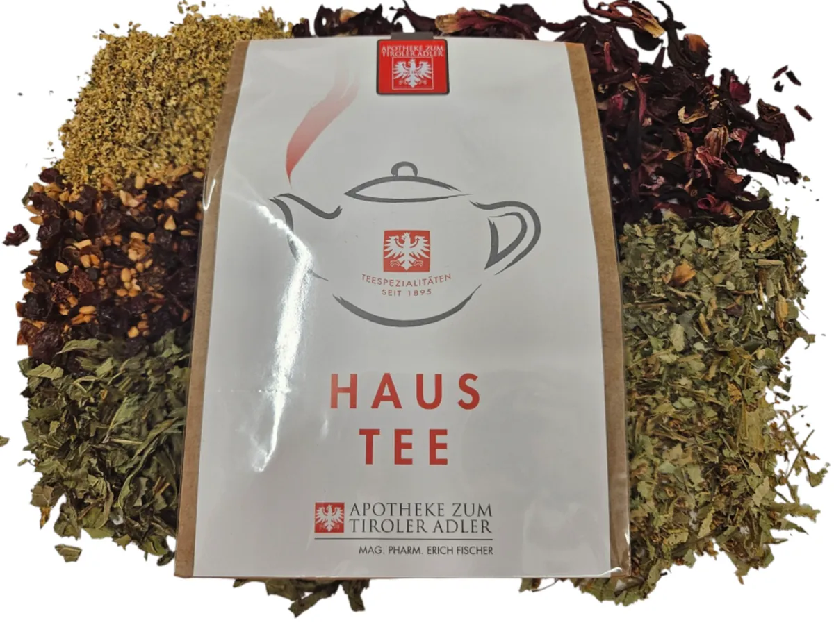 Teepackung „Haus Tee“ mit dem Logo der Apotheke zum Tiroler Adler vor Häufchen loser Teekräuter