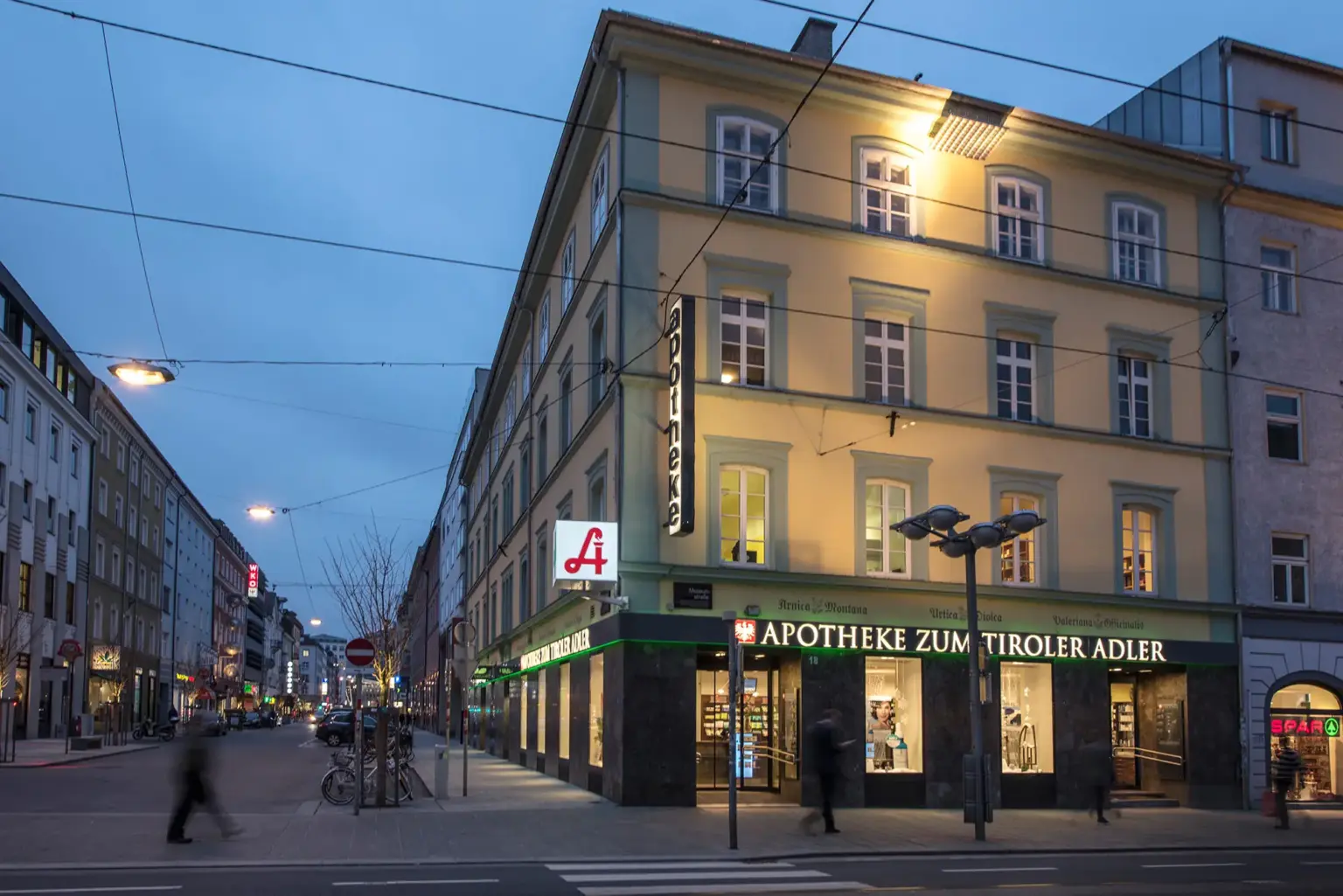 Das Eckhaus Museumstraße 18 zur blauen Stunde: beleuchtete Fassade mit Apotheken-A, senkrechtem Schriftzug „apotheke“ und „Apotheke zum Tiroler Adler“ über den Schaufenstern