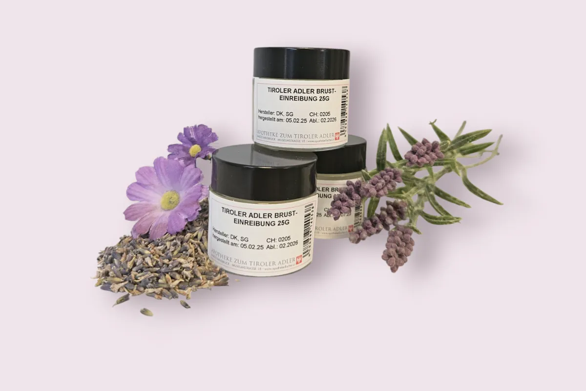 Drei Gläser mit schwarzen Deckeln und dem Etikett „Tiroler Adler Brust-Einreibung 25 g“ zwischen Lavendel und Blüten